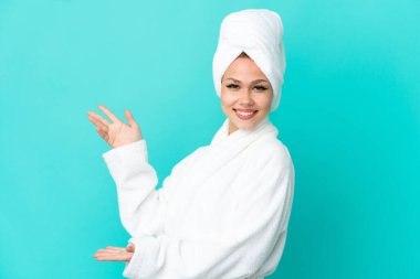 İzole edilmiş mavi arka planda bornozlu genç sarışın kız davet etmek için ellerini yana uzatıyor.