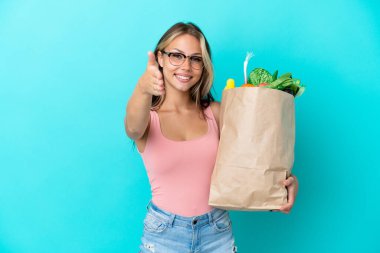 Elinde alışveriş çantası tutan genç bir Rus kadın mavi arka planda izole edilmiş ve iyi bir anlaşma için el sıkışıyor.
