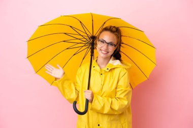 Yağmura dayanıklı paltosu ve şemsiyesi olan genç Rus kız pembe arka planda izole edilmiş ve davet edilmek için ellerini yana uzatıyor.