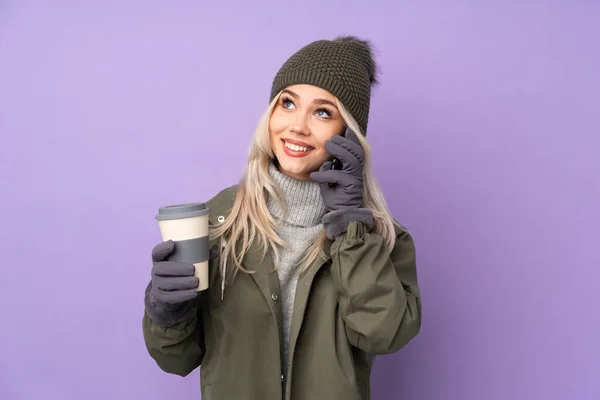 年轻的金发女郎 头戴冬帽 背景是紫色的 手里拿着咖啡和手机 — 图库照片