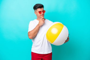 Young caucasian man holding a beach ball isolated on blue background looking to the side and smiling