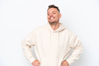 Beyaz arka planda soyutlanmış beyaz tenli yakışıklı genç adam kolları kalçasında ve gülümsüyor.