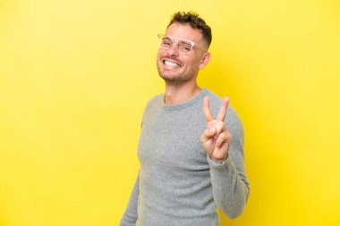 Sarı arka planda izole edilmiş yakışıklı genç adam gülümsüyor ve zafer işareti gösteriyor.