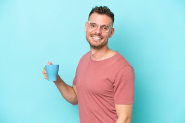Kafkasyalı genç bir adam elinde kahve fincanı tutarken mavi arka planda izole bir şekilde gülümsüyor.