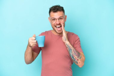 Kafkasyalı genç bir adam elinde kahve fincanı ile mavi arka planda izole bir şekilde ağzı açık bağırıyor.