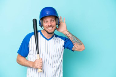 Kasklı beyzbol oyuncusu ve beysbol sopası mavi arka planda izole edilmiş bir şekilde elini kulağına koyarak bir şey dinliyor.