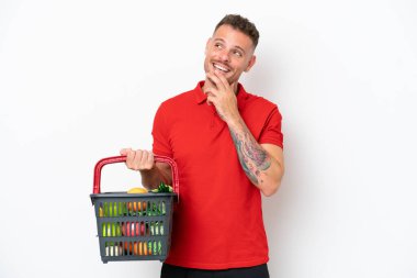 Beyaz arka planda izole edilmiş bir alışveriş sepeti taşıyan beyaz bir adam gülümserken yukarı bakıyor.