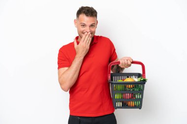 Beyaz arka planda izole edilmiş yiyecek dolu bir alışveriş sepeti tutan genç beyaz bir adam mutlu ve ağzı eliyle kaplayan bir gülümseme