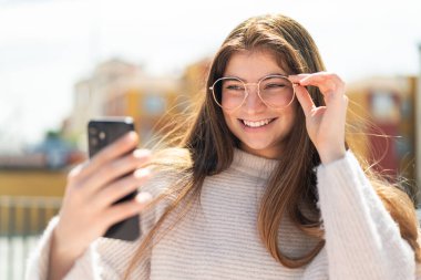 Dışarıda gözlüklü beyaz bir kadın selfie çekiyor.