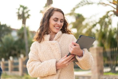 Açık havada elinde tablet tutan beyaz bir kadın, neşeli bir ifadeyle.