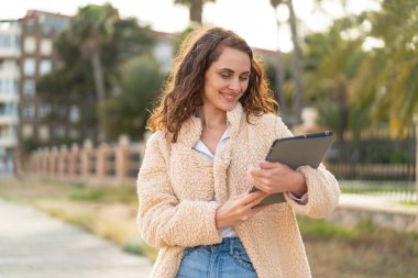 Açık havada elinde tablet tutan beyaz bir kadın, neşeli bir ifadeyle.