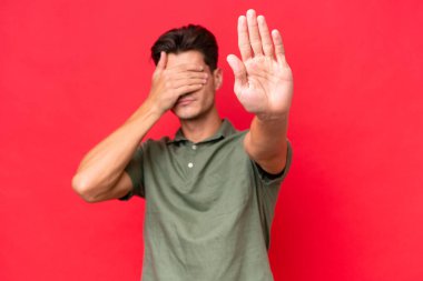 Genç beyaz yakışıklı adam kırmızı arka planda izole edilmiş. Durup jest yapıyor ve yüzünü kapatıyor.