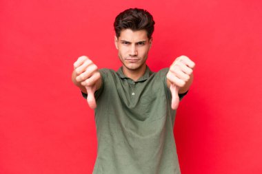 Kızıl arka planda izole edilmiş, iki eliyle baş parmağını gösteren beyaz, yakışıklı genç bir adam.