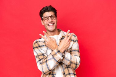 Kızıl arka planda izole edilmiş genç beyaz adam gülümsüyor ve zafer işareti gösteriyor.