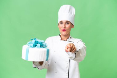 Orta yaşlı beyaz bir kadın, büyük bir pastayla soyutlanmış arka planı şaşırmış ve ön tarafı işaret ediyor.