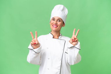 Orta yaşlı aşçı kadın izole edilmiş arka planda zafer işaretini iki eliyle gösteriyor.