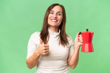 Orta yaşlı beyaz bir kadın kahve makinesini izole edilmiş arka planda tutuyor. Başparmakları havada çünkü iyi bir şey oldu.