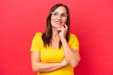 Orta yaşlı beyaz kadın, kırmızı arka planda izole edilmiş, yukarı bakarken bir fikir düşünüyor.