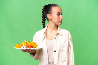 İzole edilmiş arka planda waffle tutan genç bir Arap kadın yan tarafa bakıyor.