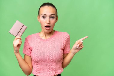 Soyutlanmış arka planda cüzdanını tutan genç bir Arap kadın şaşırmış ve işaret ediyor.