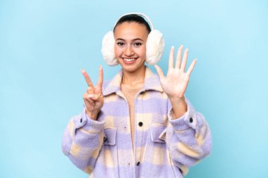 Mavi arka planda izole edilmiş kışlık kuklalar giyen genç Arap kadın parmaklarıyla yediyi sayıyor.