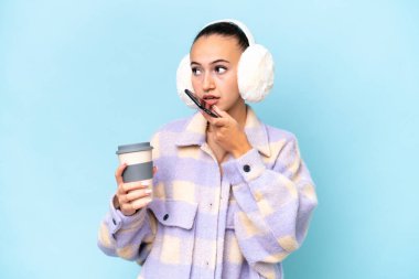 Mavi arka planda izole edilmiş kışlık kuklalar takan genç bir Arap kadın elinde kahve ve bir cep telefonu tutuyor.