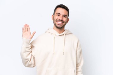 Beyaz arka planda izole edilmiş genç beyaz bir adam mutlu bir ifadeyle selamlıyor.