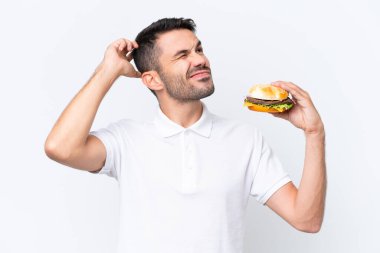 Genç, yakışıklı, beyaz bir adam arka planda bir burger tutuyor. Şüpheleri var ve kafası karışık bir yüz ifadesi var.
