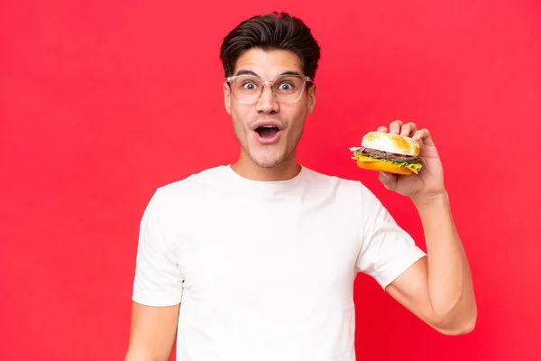 Młody Biały Mężczyzna Trzymając Hamburgera Odizolowany Czerwonym Tle Zaskoczeniem Wstrząśnięty — Zdjęcie stockowe