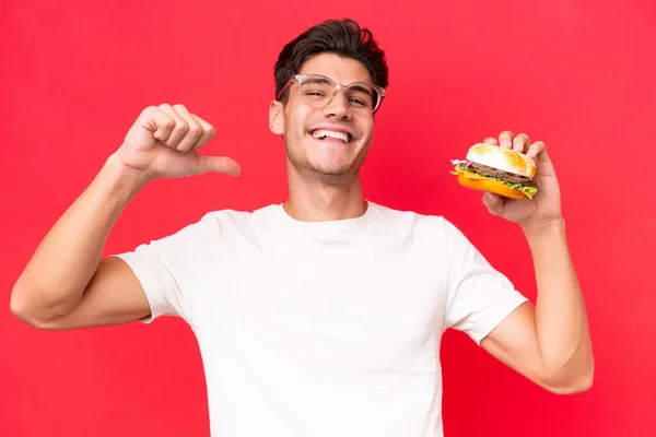 Jonge Blanke Man Met Een Hamburger Geïsoleerd Rode Achtergrond Trots — Stockfoto