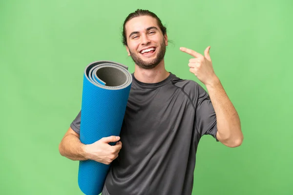 年轻的运动员一边带着垫子去上瑜伽课 一边用大拇指举手表决孤立的背景 — 图库照片