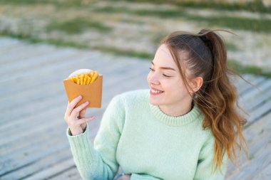 Genç ve güzel bir kız açık havada mutlu bir ifadeyle patates kızartması tutuyor.