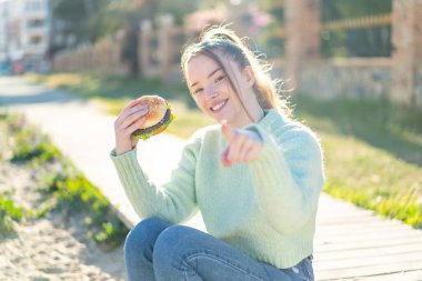 Genç ve güzel bir kız açık havada bir burger tutuyor. Kendini beğenmiş bir ifadeyle seni işaret ediyor.