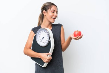 Genç, güzel Uruguaylı bir kadın beyaz arka planda izole edilmiş. Elma ararken elinde bir tartı makinesi tutuyor.