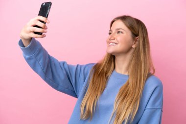 Pembe arka planda izole edilmiş genç sarışın kadın cep telefonuyla selfie çekiyor.