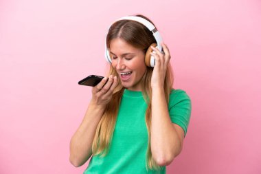 Pembe arka planda izole edilmiş genç sarışın bir kadın cep telefonuyla müzik dinliyor ve şarkı söylüyor.