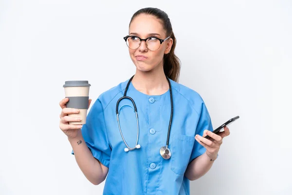 年轻的高加索外科医生女医生被隔离在白色背景下 一边拿着咖啡一边想着什么 一边拿着手机走了 — 图库照片
