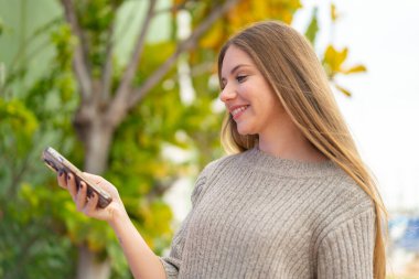 Açık havada cep telefonu kullanan güzel, sarışın bir kadın. Mutlu bir ifadeyle.