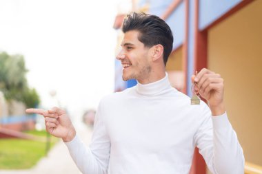 Kafkasyalı genç bir adam evin anahtarlarını elinde tutuyor ve bir ürünü sunmak için yan tarafı işaret ediyor.