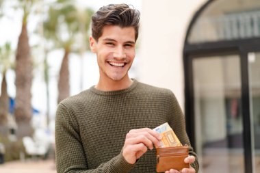 Açık havada genç beyaz bir adam elinde cüzdanla mutlu bir ifadeyle.