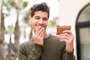 Açık havada genç beyaz bir adam bir cüzdan tutuyor ve ona bakıyor.