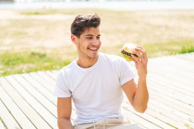 Genç beyaz bir adam açık havada elinde mutlu bir ifadeyle bir burger tutuyor.