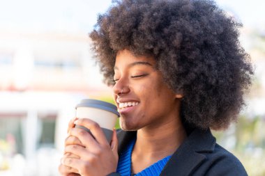 Afro-Amerikalı bir kız açık havada elinde neşeli bir ifadeyle kahve tutuyor.