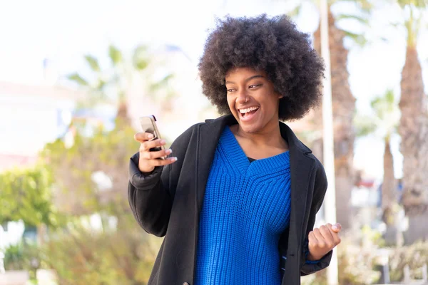 African American Girl Outdoors Using Mobile Phone Doing Victory Gesture — Stock Photo, Image