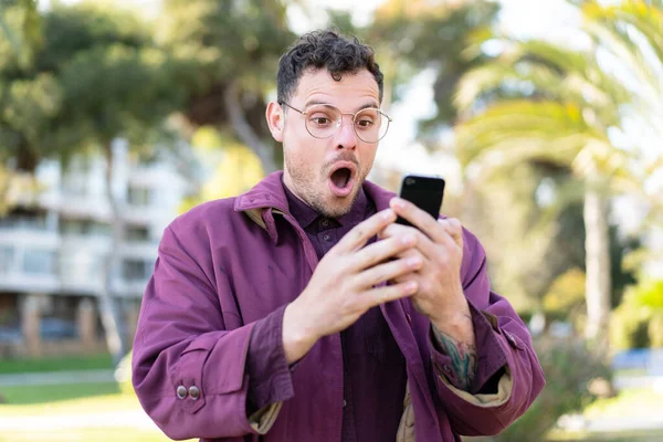 Young Caucasian Man Outdoors Looking Camera While Using Mobile Surprised — Stock Photo, Image