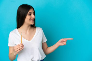 Mavi arka planda dişlerini fırçalayan genç beyaz kadın bir ürünü sunmak için yan tarafı işaret ediyor.