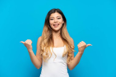 Mavi arka planda izole edilmiş genç bir Rus kızı. Baş parmakları havada ve gülümsüyor.