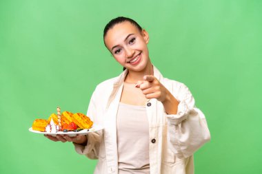 İzole edilmiş arka planda waffle tutan genç bir Arap kadını mutlu bir ifadeyle ön tarafı işaret ediyor.