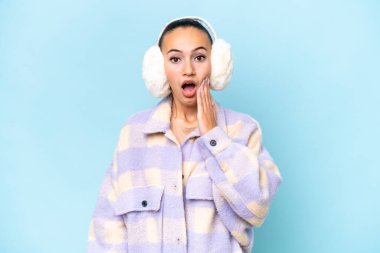 Mavi arka planda, şaşırmış ve şok olmuş yüz ifadesiyle izole edilmiş, kış maskeleri takan genç bir Arap kadın.