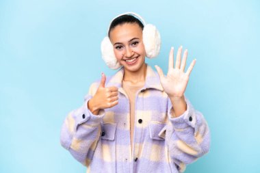 Mavi arka planda izole edilmiş kış pantolonu giyen genç Arap kadın parmaklarıyla altısını sayıyor.